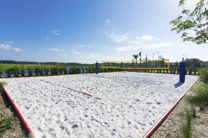 A sandy beach volleyball court with a net, surrounded by greenery and a clear sky.