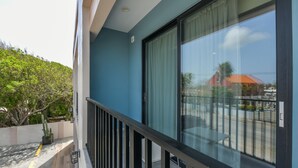 Un balcón con puerta de vidrio, baranda metálica y vista a un edificio y árboles.