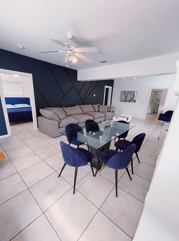 A modern living room with a glass dining table and blue chairs, a dark accent wall, and a ceiling fan.