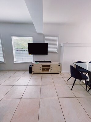 A modern living room with a flat-screen TV mounted on the wall, a wooden cabinet, and a dining table with chairs.