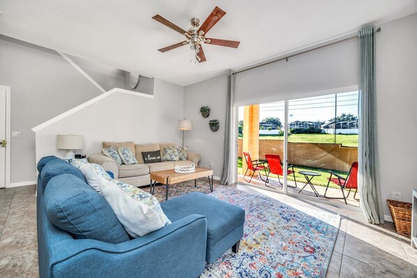 A modern living room with a blue sofa, a wooden coffee table, and a ceiling fan. There is a large sliding glass door leading to an outdoor patio with red chairs and a table.