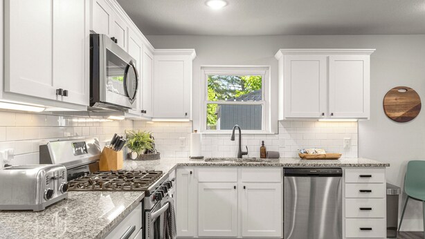 Una cocina con armarios blancos, electrodomésticos de acero inoxidable, un ventanal con vistas y una tabla de cortar de madera en la pared.