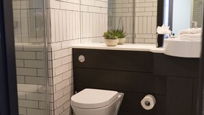A bathroom with white tiled walls, a white toilet, a dark sink vanity, and a glass shower enclosure. - Dublin One Hotel (Dublin)