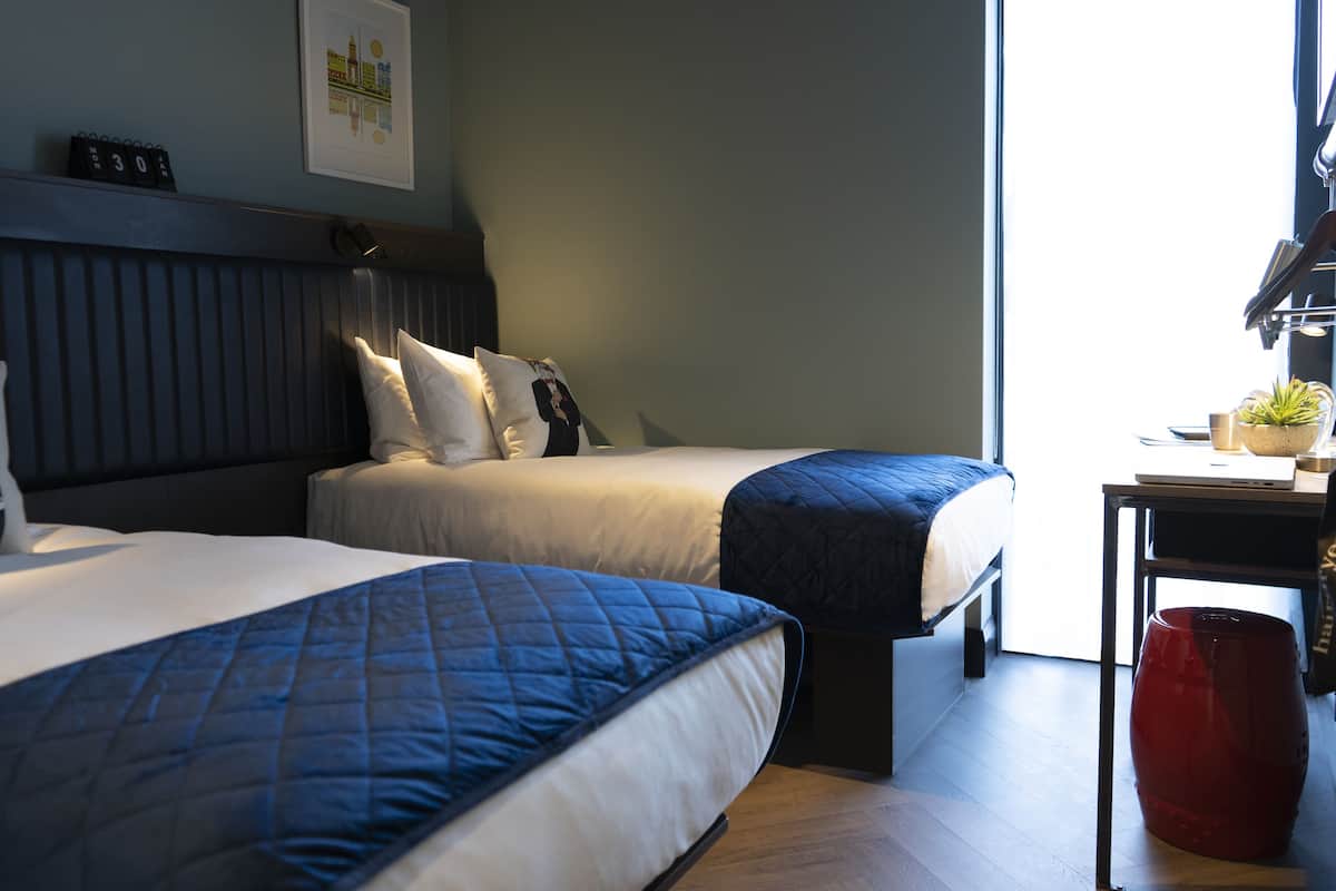 Habitación de hotel con dos camas, un escritorio con computadora, un cesto de basura rojo y una ventana con luz natural.