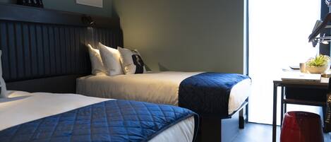 A hotel room with two beds, a desk with a computer, a red trash bin, and a window with natural light.