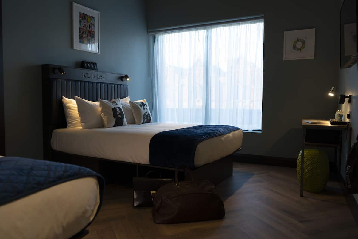 Un dormitorio con cama, mesita de noche, lámpara y ventana con cortinas translúcidas.