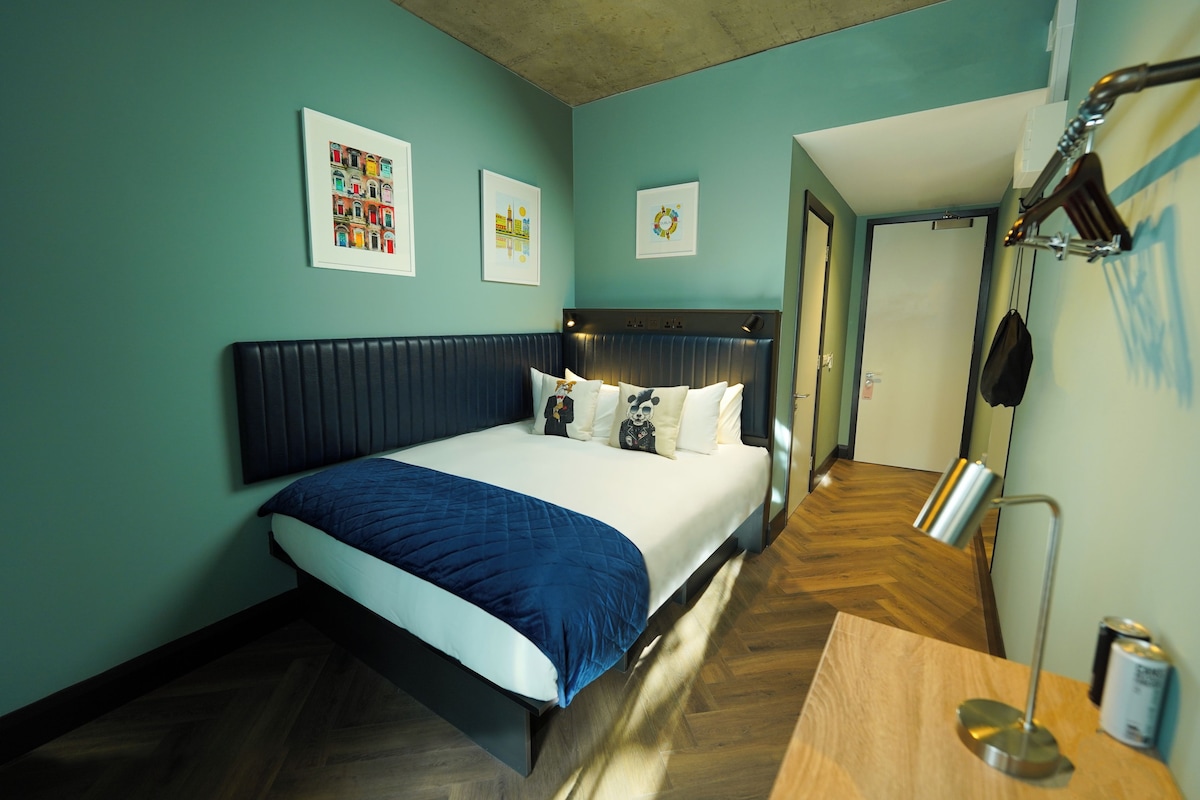 A hotel room with a bed, a desk, and a chair. There are framed pictures on the wall and a door leading to another room.