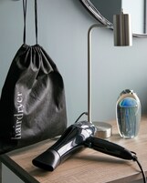 Un secador de pelo, una bolsa de tela negra con cordón y un florero de vidrio con diseño azul y verde sobre una superficie de madera.