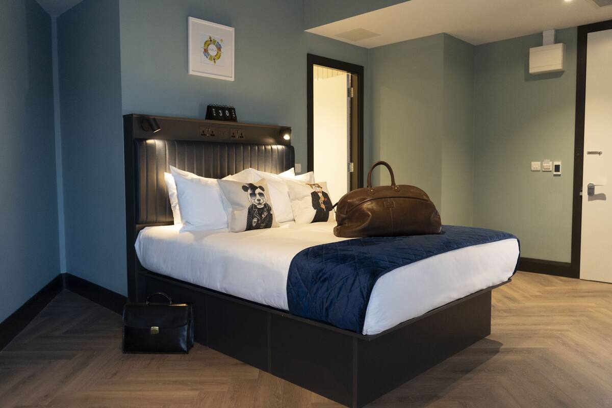 Un dormitorio moderno con una cama grande, cabecera con iluminación integrada, mesita de noche con un bolso y una puerta que da a otra habitación.