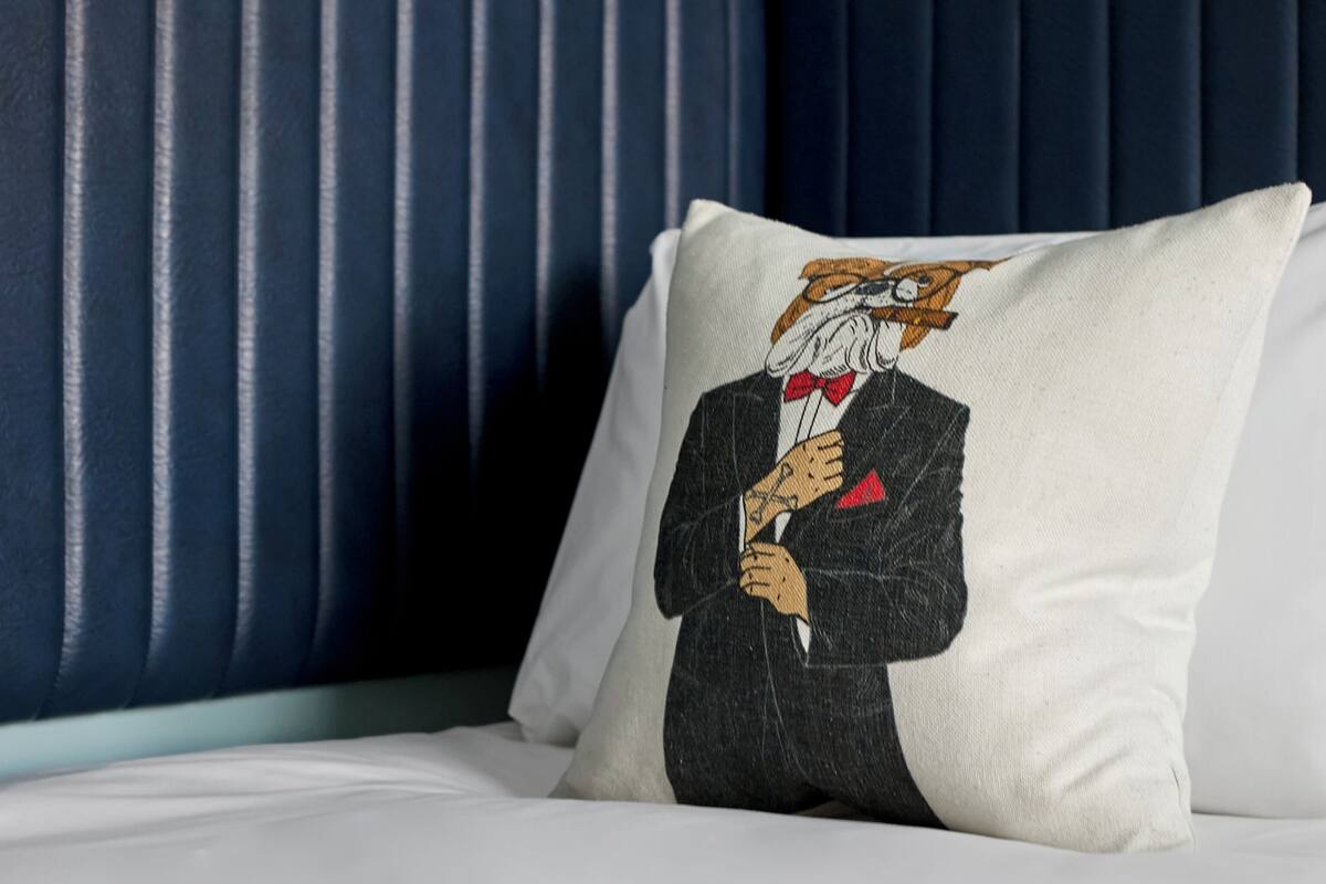 Un almohadón con diseño gráfico sobre una cama con cabecera tapizada en azul.