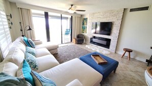 A living room with a sofa, ottoman, and a flat-screen TV mounted on a brick wall. - Sand Dollar 101 (St. Augustine)