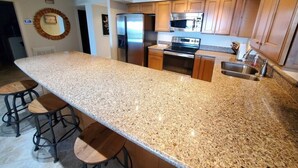 A kitchen with granite countertops, stainless steel appliances, wooden cabinets, and a large island with bar stools. - Sand Dollar 101 (St. Augustine)