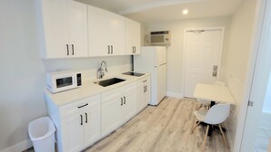 Cocina compacta con muebles blancos, microondas, pileta y refrigerador. Hay un bote de basura pequeño y una silla cerca de la puerta.