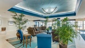 Un hall d’entrée spacieux, aux couleurs bleu et blanc, comprenant un grand espace salon avec des canapés et des fauteuils, plusieurs plantes en pot et un design de plafond original.
