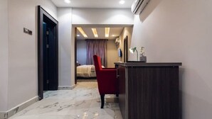 Un pasillo de hotel con piso de mármol, una silla roja, un escritorio y una cama al fondo.
