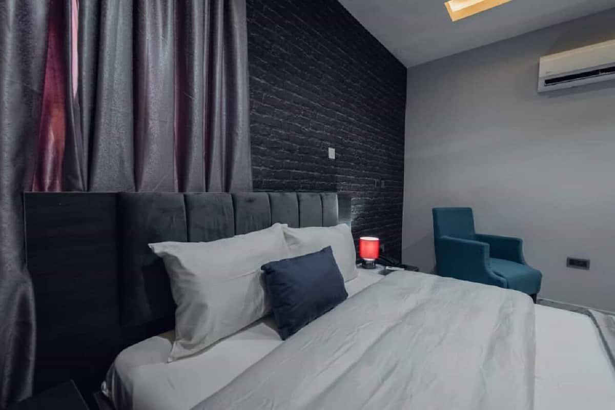 A modern hotel room with a dark wood headboard, a bed with white and dark blue bedding, a red table lamp, and a blue armchair.