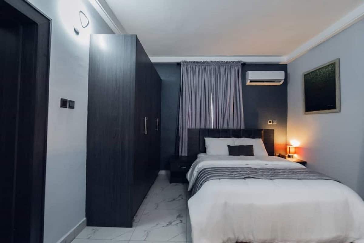 A bedroom with a bed, a wardrobe, a nightstand, a lamp, and an air conditioning unit.