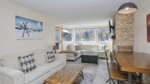 A modern living room with a sofa, coffee table, and dining table. There are large windows with a view of snow-covered mountains. - Convenient 1 Bedroom East Vail Condo by Gorecreek Rentals (Vail)
