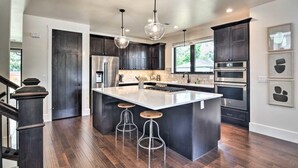 Une cuisine moderne avec des meubles de couleur sombre, un grand îlot central doté de tabourets, des appareils électroménagers en acier inoxydable et un éclairage suspendu.