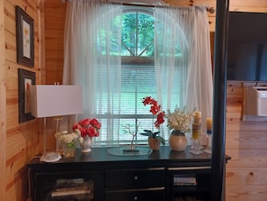 Interior d'una cabana de fusta amb un escriptori, quadres i una finestra amb cortines translúcides.