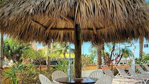 A thatched umbrella table with white plastic chairs, set on a paved patio with palm trees in the background. - Satellite Motel - License to Chill 115 (Treasure Island)