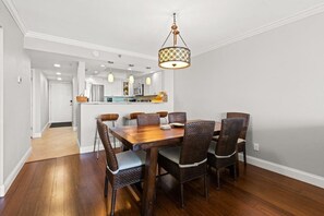 A dining area with a wooden table and wicker chairs, a hanging light fixture, and a kitchen in the background.