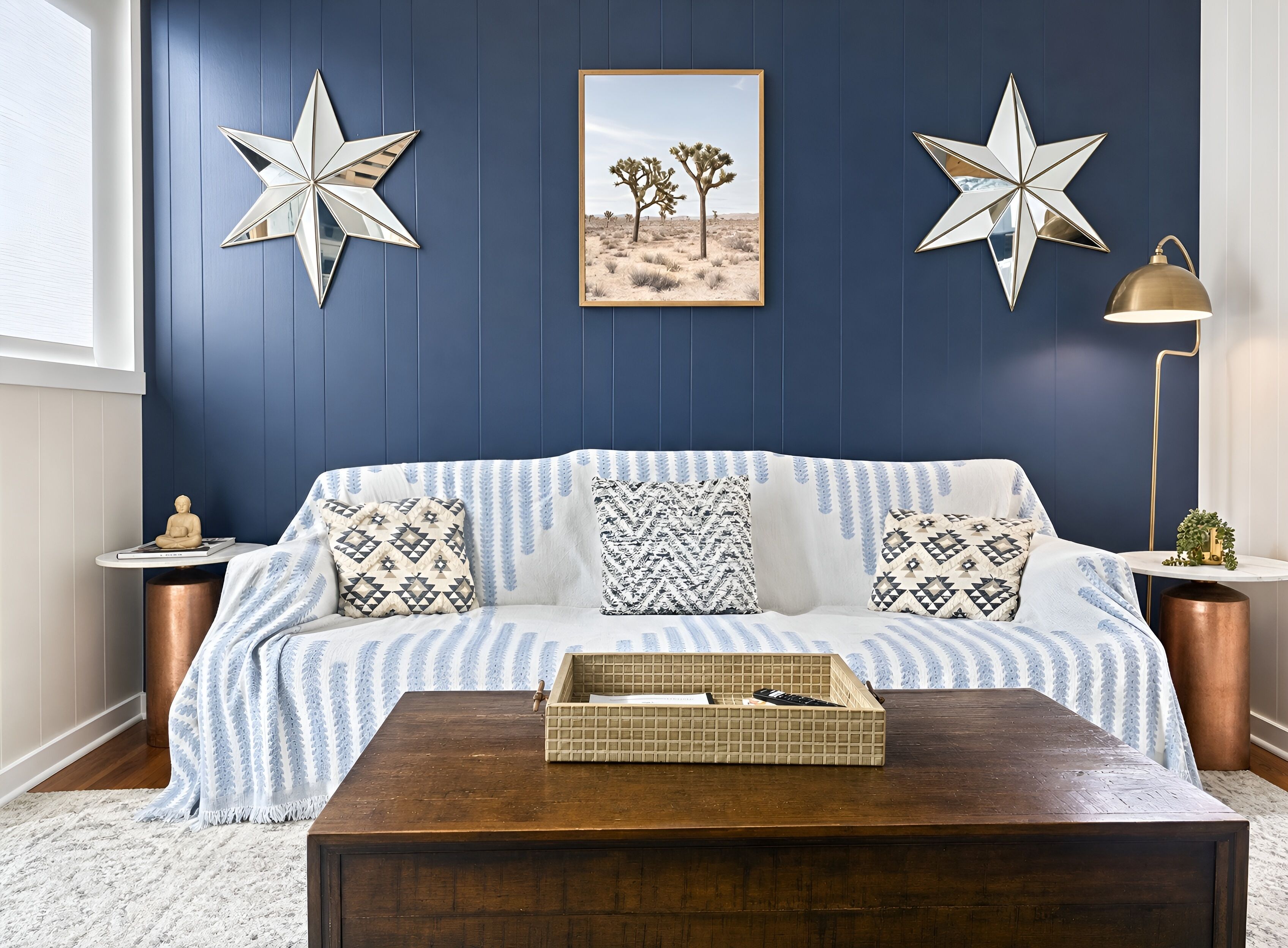 A blue-walled room with a white sofa, patterned cushions, a star decoration, a framed desert landscape, a floor lamp, and a wooden coffee table.