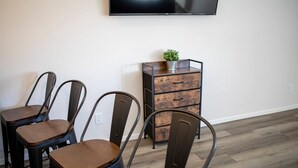 Un comedor con cuatro sillas de madera y una mesa de metal, un armario de madera con cajones y un televisor montado en la pared.