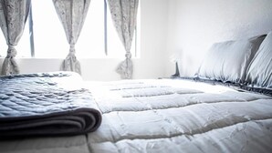 Una cama bien hecha con una manta doblada, una mesita de noche con una lámpara y una ventana con cortinas.