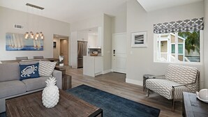A modern living room with a sofa, coffee table, and a chair. There is a kitchen area in the background with a refrigerator and cabinets. - Chic Upstairs Condo Steps from Strands Beach (Dana Point)