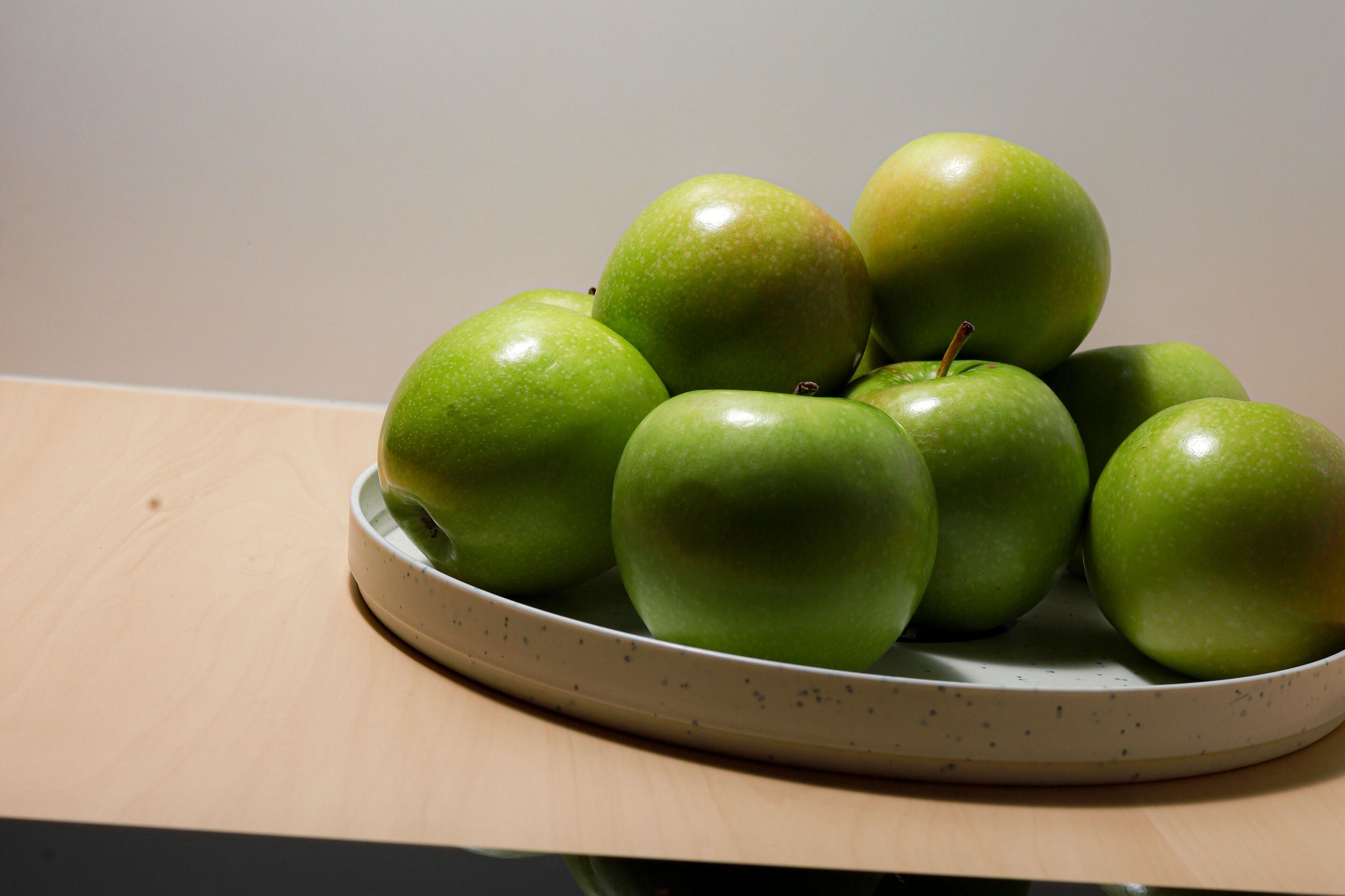 A ceramic plate with several green apples on a wooden surface.