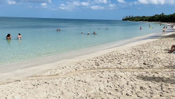 Une plage à l’eau turquoise et limpide, avec un rivage sablonneux, où quelques personnes se baignent et se promènent.
