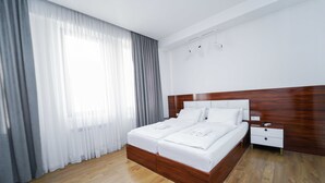Ein modernes Hotelzimmer mit einem hölzernen Bett, weißen Bettwäsche, einem Nachttisch aus Holz und einem großen Fenster mit durchscheinenden Vorhängen.