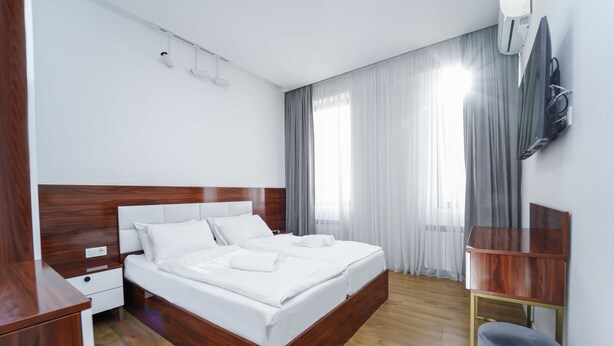 Ein modernes Hotelzimmer mit einem hölzernen Bett, einem kleinen Nachttisch, einem eingelassenen Fernseher und einem grau gepolsterten Hocker.