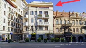 Ein mehrstöckiges Gebäude mit einem Laden im Erdgeschoss, Balkonen mit Pflanzen und einer Fußgängerquerung.