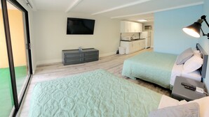 A bedroom with two beds, a TV, a desk with a lamp, and a kitchenette in the background.