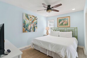 A bedroom with a bed, a ceiling fan, a television, artwork, and a window with blinds. - One Lot off the Beach, a Sophisticated Chic East Beach Retreat w Heated Pool (Saint Simons Island)