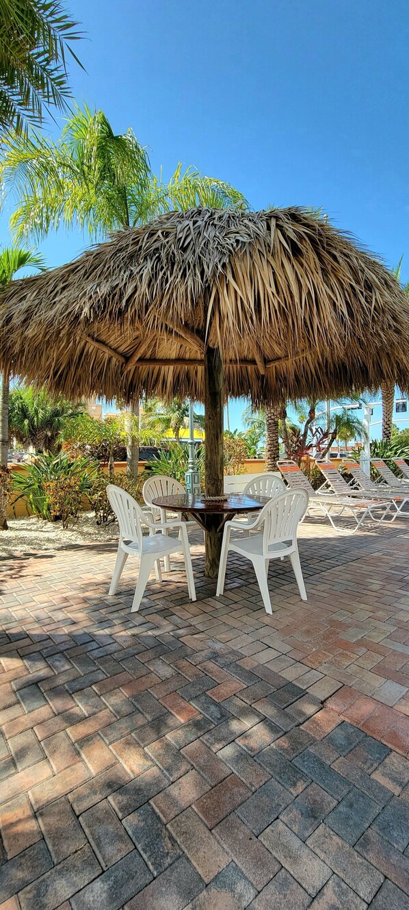 Une table parasol à toit de chaume, entourée de chaises en plastique blanc, installée sur une terrasse pavée avec des palmiers en arrière-plan.
