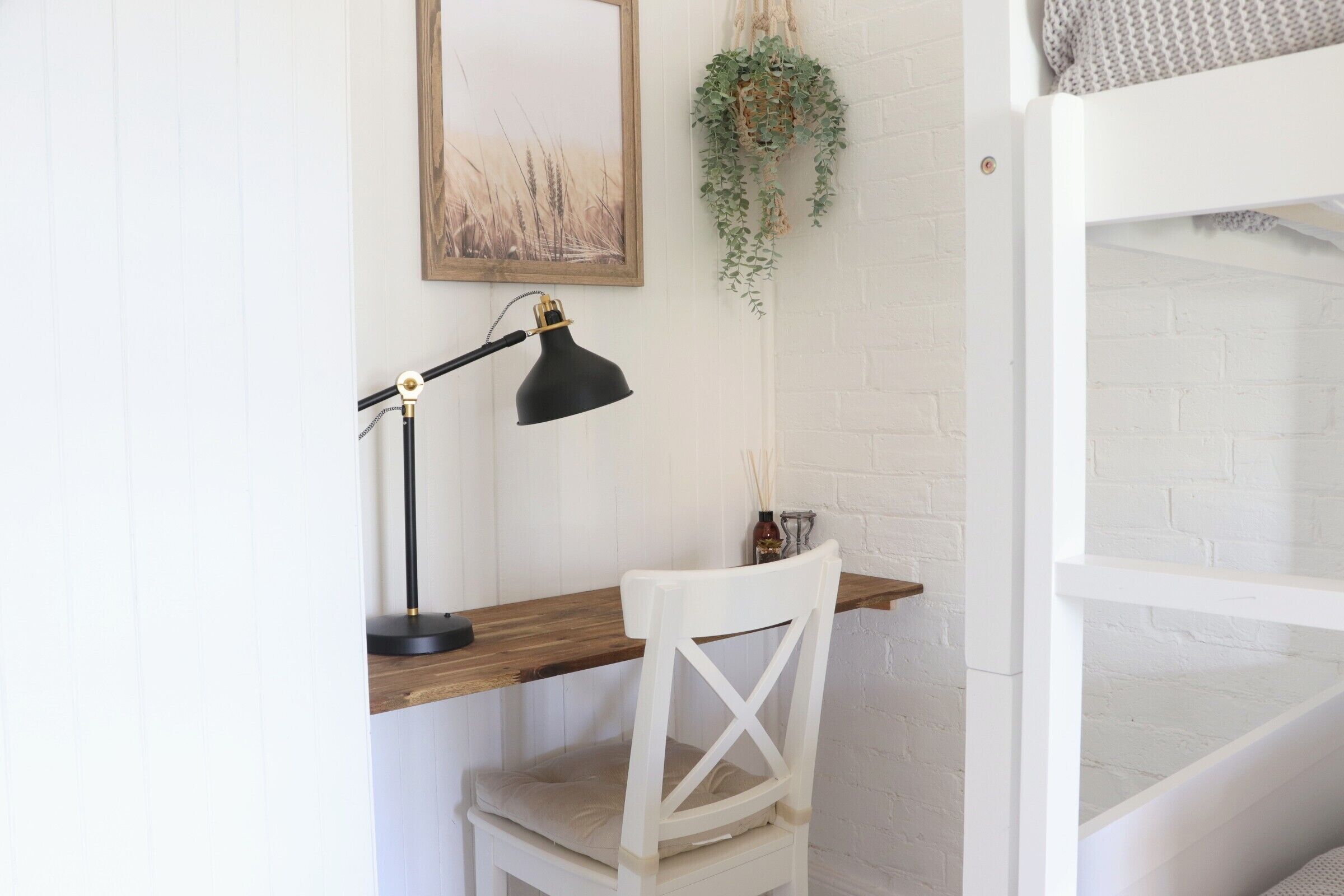 Une chaise blanche, un bureau en bois, une lampe de bureau noire, un tableau encadré accroché au mur et une plante suspendue.