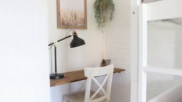 Une chaise blanche, un bureau en bois, une lampe de bureau noire, un tableau encadré accroché au mur et une plante suspendue.