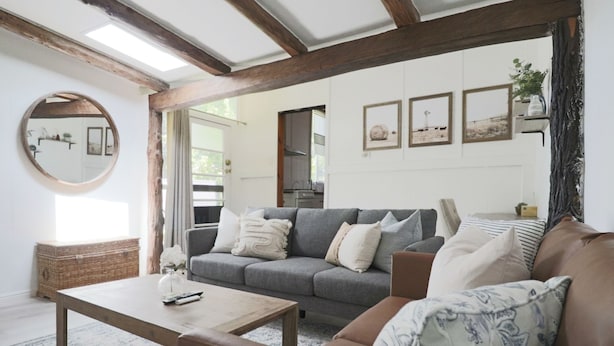 Un salon avec un canapé gris, un divan en cuir marron, une table basse en bois et un miroir rond. Des tableaux encadrés ornent le mur et une plante est posée sur une étagère.