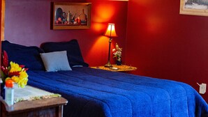 Ein Schlafzimmer mit roter Wand, blauer Bettdecke, einem Nachttisch mit Lampe und einem Bild an der Wand.