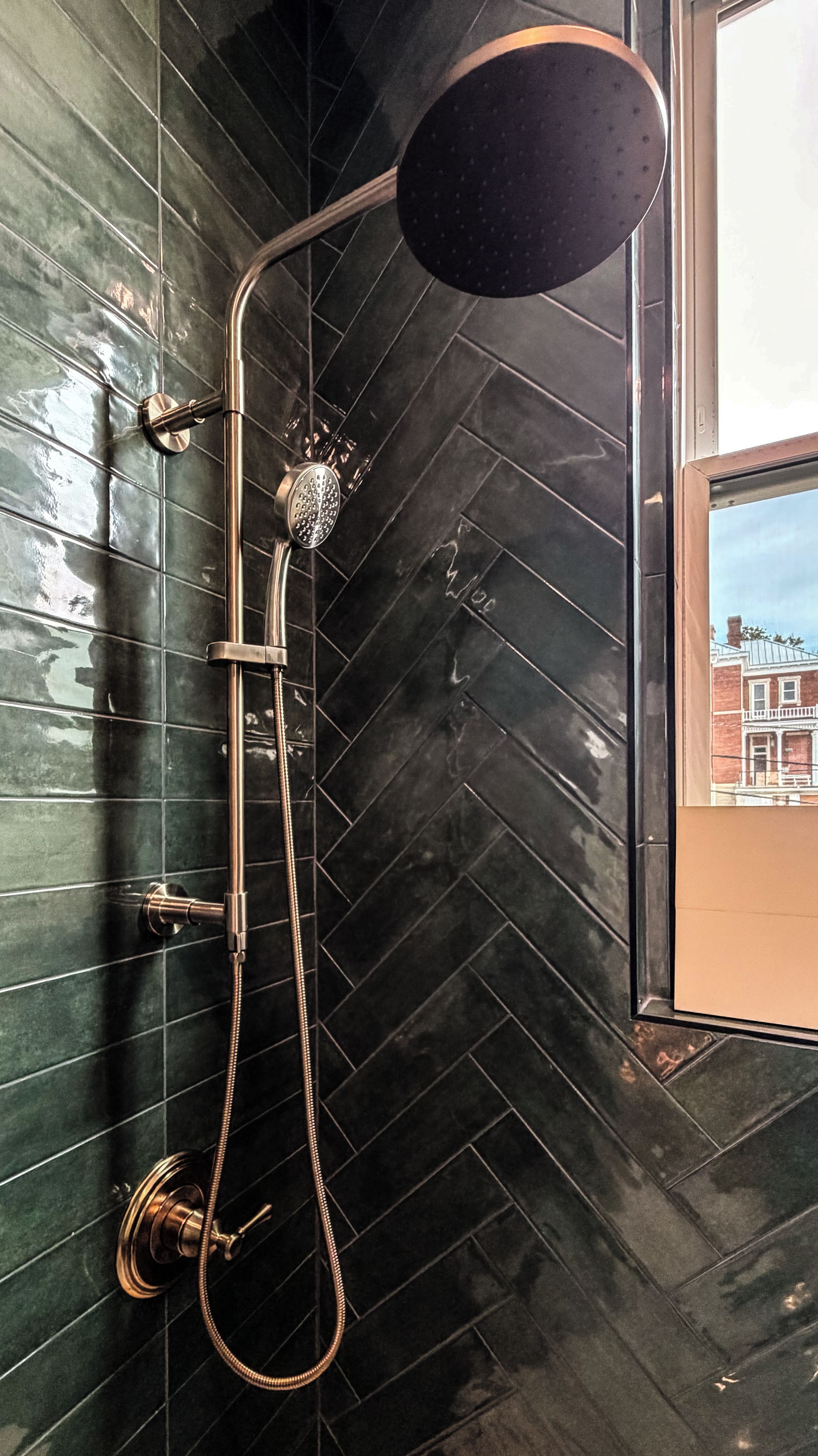 A shower with a handheld showerhead and a wall-mounted rain showerhead, both with metallic finishes, against a dark tiled wall.