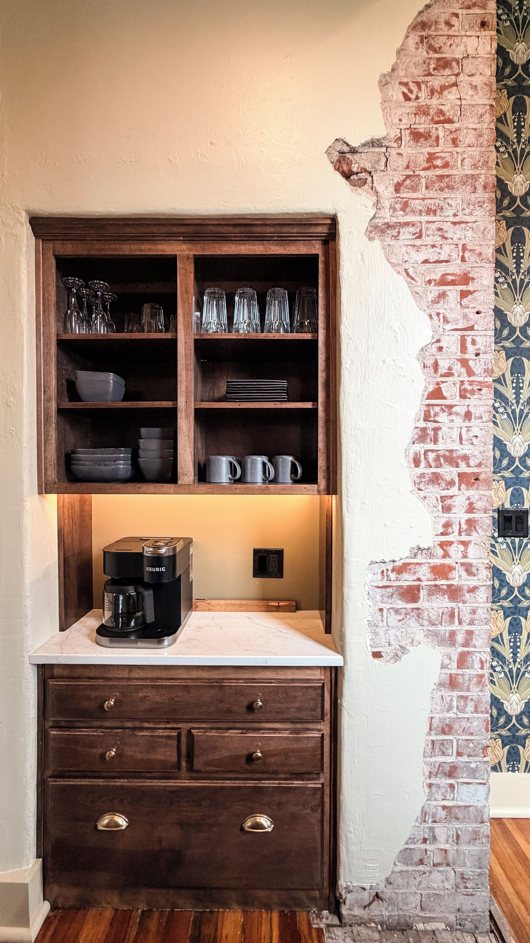 Une cuisine avec une machine à café sur le plan de travail, un meuble en bois contenant de la vaisselle et un mur décoratif orné d’un motif en briques.
