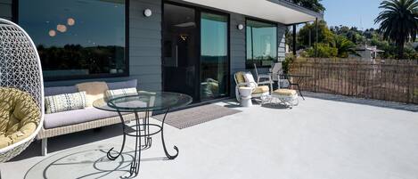 A modern outdoor patio with a glass table, wicker sofa, and white chairs.