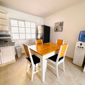 A dining area with a wooden table and chairs, a refrigerator, a water dispenser, and a window with blinds.