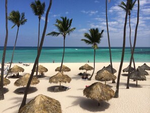 Une plage bordée de parasols en chaume et de palmiers, un rivage sablonneux et une mer d’un bleu limpide.