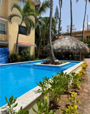 Una piscina con una estructura de techo de paja, rodeada de palmeras y un edificio con techo de tejas.