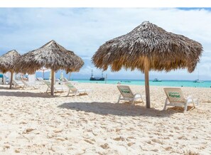 Una playa con sombrillas de paja y sillones blancos.