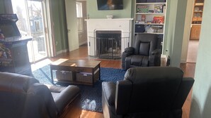 Een woonkamer met een open haard, leren fauteuils, een bijzettafeltje en een boekenkast met gezelschapsspellen.